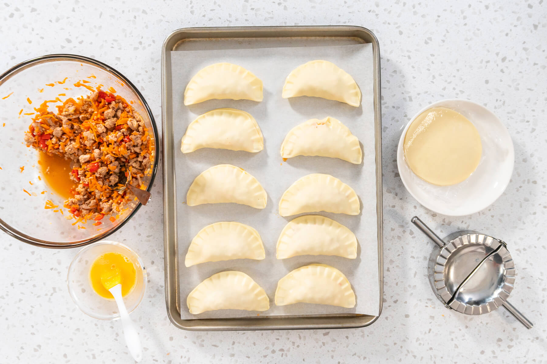 Blech mit zehn ungebackenen Puten-Empanadas, daneben die Schüssel mit restlicher Füllung und Saft am Boden, Teigscheiben, verquirltes Ei und Teigtaschenpresse