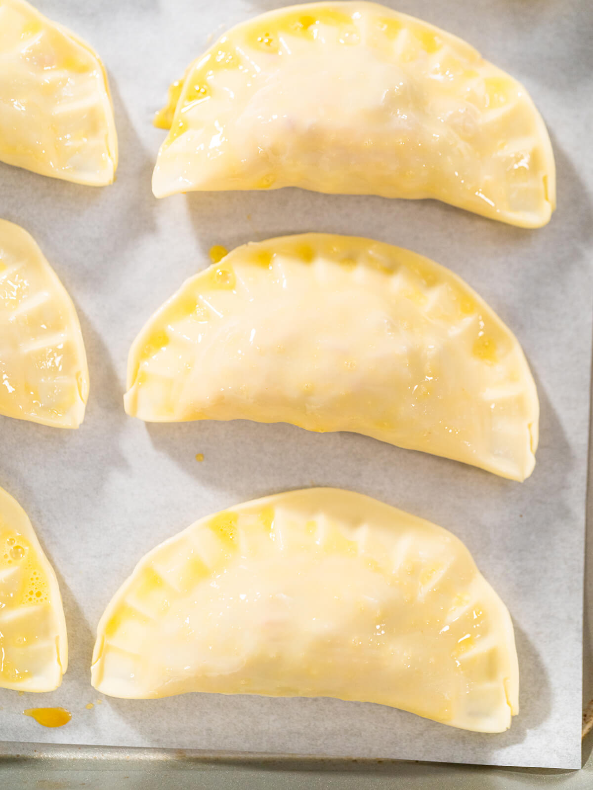 Nahaufnahme von drei mit Ei bestrichenen ungebackenen Puten-Empanadas mit geriffeltem Rand auf Backpapier