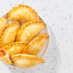 Gebackene Puten-Empanadas auf einem weißen Teller, eine aufgebrochen mit Blick auf die Füllung aus Putenhack, Karotte und Paprika