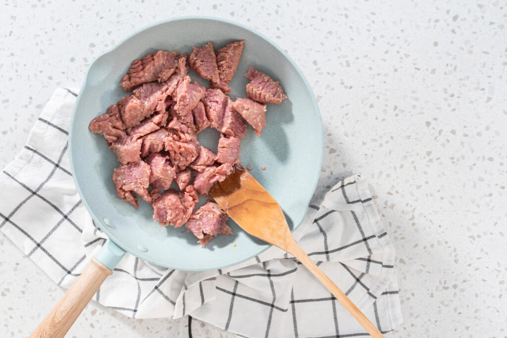 Rohes Putenhackfleisch in Stücken in einer hellblauen Pfanne mit Holzwender