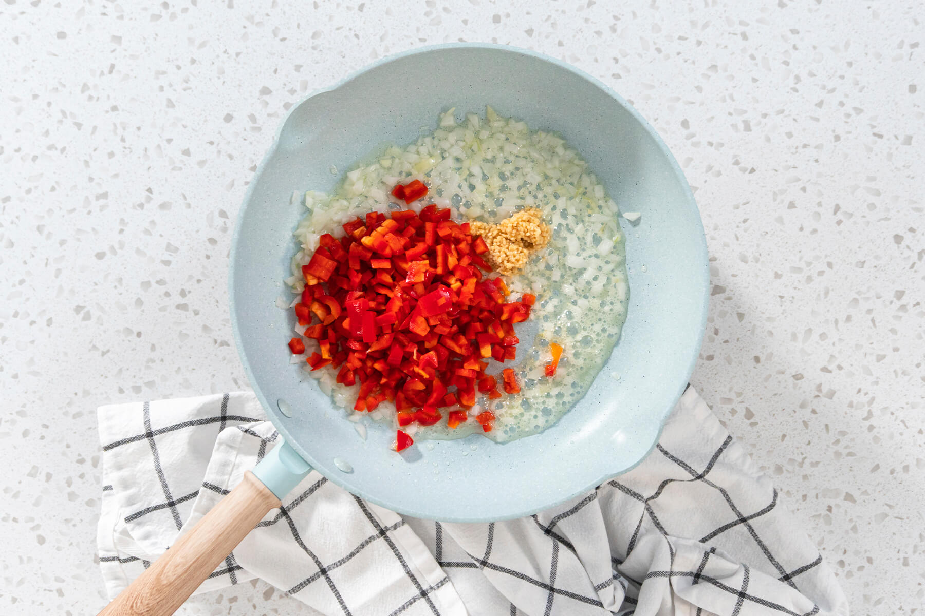 Hellblaue Pfanne mit glasigen Zwiebelwürfeln, gehacktem Knoblauch und fein gewürfelter roter Paprika in Olivenöl