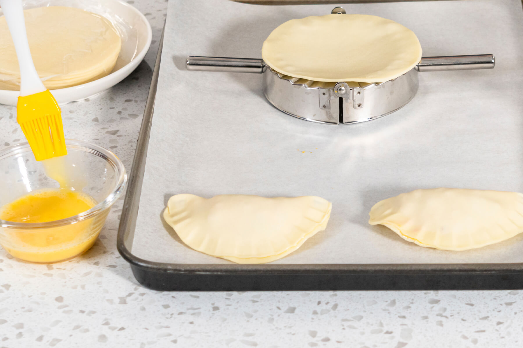 Runde Teigscheibe auf einer geschlossenen Teigtaschenpresse, daneben verquirltes Ei mit Backpinsel und zwei geformte Empanadas auf dem Blech