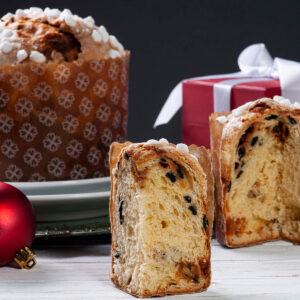 Hoher Panettone in gemusterter Papierform mit Hagelzucker auf der Kuppe, davor zwei aufgeschnittene Hälften mit dunklen Schokoladenstücken in der Krume