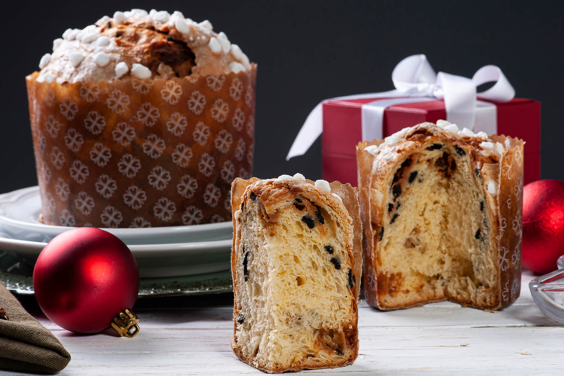 Panettone in gemusterter Papierform mit Hagelzucker, davor zwei aufgeschnittene Hälften mit dunklen Schokoladenstücken und roten Weihnachtskugeln