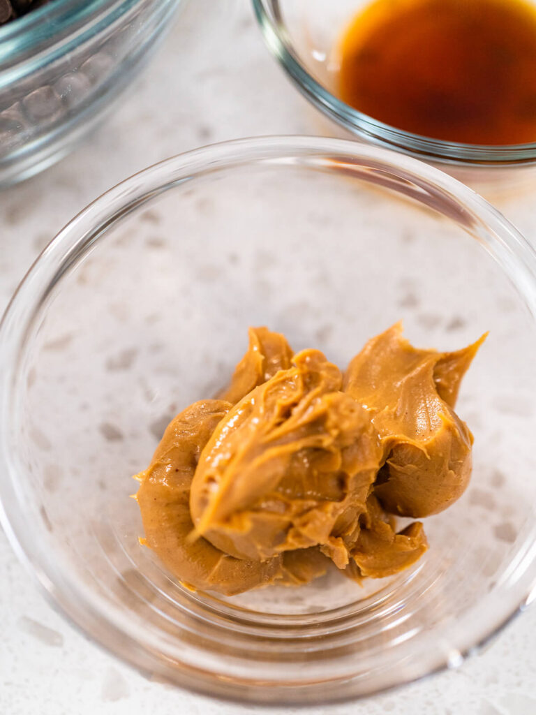 Zimmerwarme Erdnussbutter in einer kleinen Glasschale, daneben dunkle Schokoladendrops