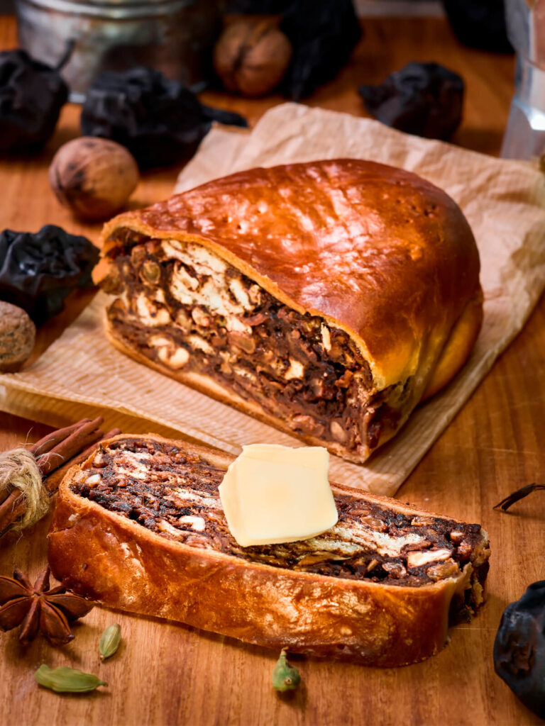 Angeschnittener Birnbrot-Laib und eine dicke Scheibe mit Butter, dahinter Zimtstangen und Sternanis