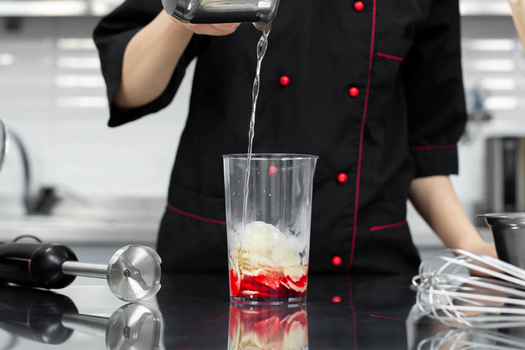 Heißer Zuckersirup wird im Mixbecher über weiße Schokolade und rote Lebensmittelfarbe gegossen