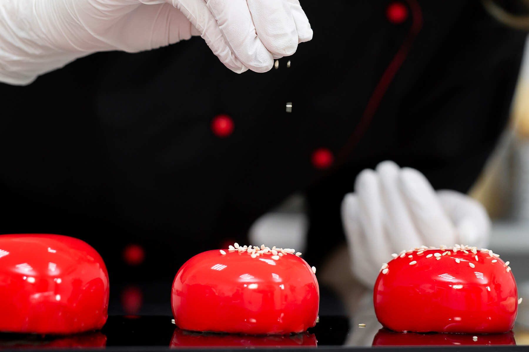 Heller Sesam wird von oben über glasierte Törtchen auf einer schwarzen Platte gestreut