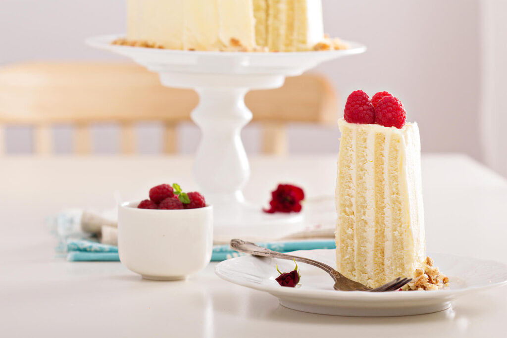 Stück vertikale Biskuittorte mit senkrechten Biskuitstreifen und Himbeeren, dahinter die angeschnittene Torte