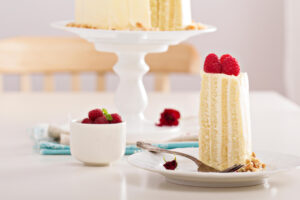 Stück vertikale Biskuittorte mit senkrechten Biskuitstreifen und Himbeeren, dahinter die angeschnittene Torte