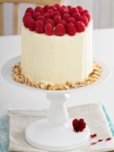 Hohe vertikale Biskuittorte mit heller Frischkäsecreme und dicht gesetzten Himbeeren auf einer weißen Tortenplatte