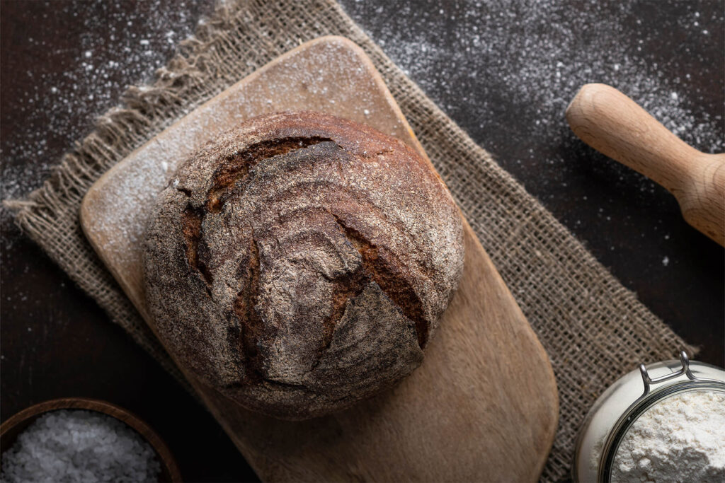 Runder Laib Vollkorn-Kartoffelbrot von oben auf einem bemehlten Holzbrett mit Nudelholz und Mehlglas