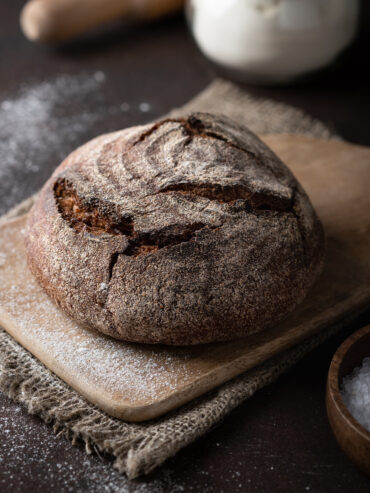Runder Laib Vollkorn-Kartoffelbrot mit dunkler aufgerissener Kruste auf einem bemehlten Holzbrett