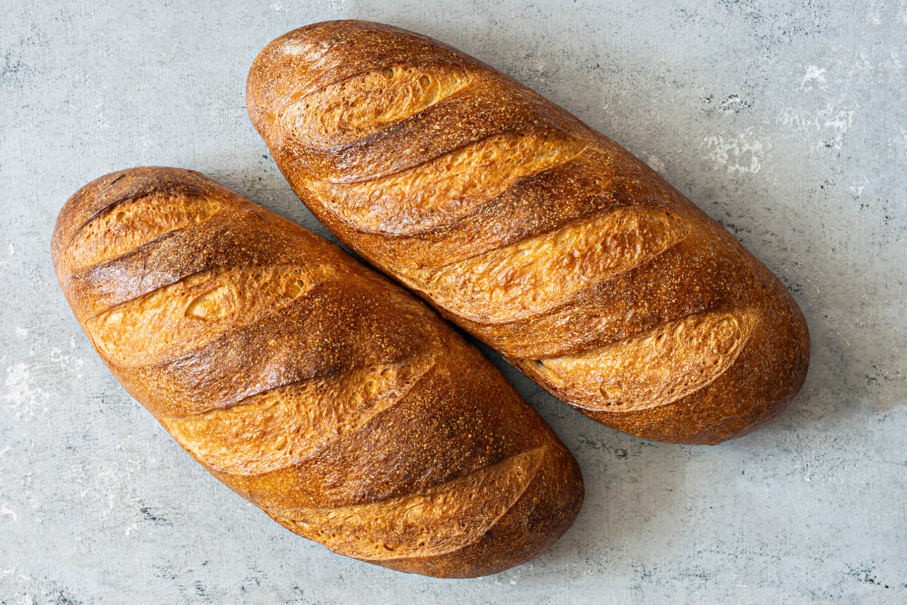 Zwei goldbraune Laibe Weißbrot aus Sauerteig-Resten nebeneinander mit je fünf Schrägschnitten
