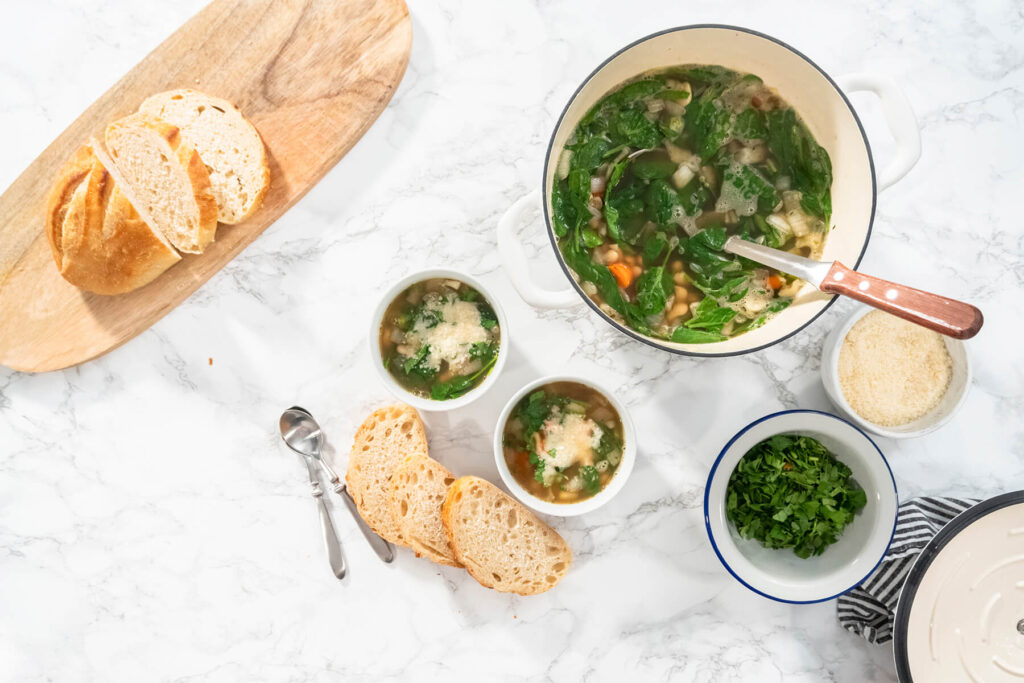 Zwei weiße Schalen italienische weiße Bohnensuppe mit Spinat und geriebenem Parmesan neben dem Topf und aufgeschnittenem Weißbrot