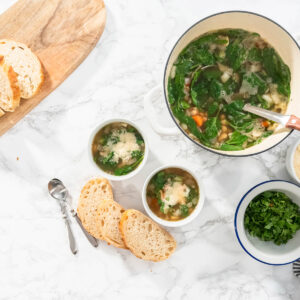 Zwei weiße Schalen italienische weiße Bohnensuppe mit Spinat und geriebenem Parmesan neben dem Topf und aufgeschnittenem Weißbrot