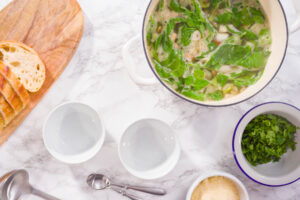 Fertige weiße Bohnensuppe mit Spinat im Topf, daneben leere Schalen, Petersilie, geriebener Parmesan und Weißbrot