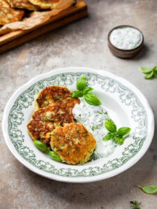 Zucchini-Fritter mit Parmesan und Tzatziki, serviert auf einem Teller mit Basilikum