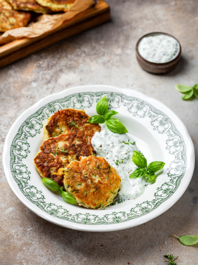 Zucchini-Fritter mit Parmesan und Tzatziki, serviert auf einem Teller mit Basilikum