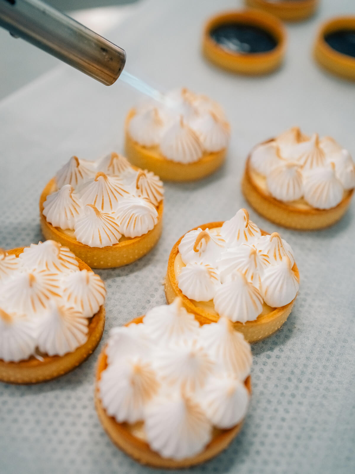 Ein Küchenbrenner bräunt aus kurzer Entfernung die Spitzen einer aus Tupfen aufgespritzten Baiserhaube auf einem Tartelette