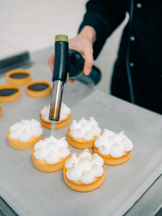 Eine Hand führt einen Küchenbrenner über mehrere Tartelettes mit weißer Baiserhaube auf einem Backblech