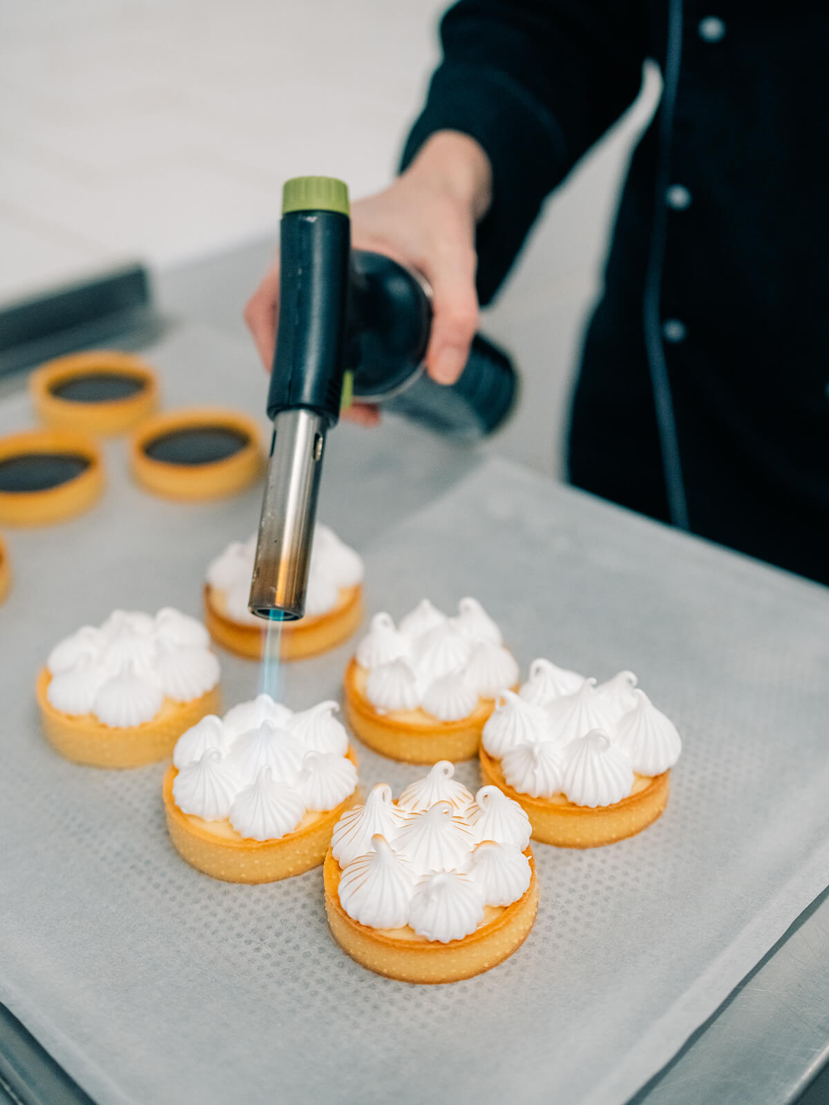 Eine Hand führt einen Küchenbrenner über mehrere Tartelettes mit weißer Baiserhaube auf einem Backblech