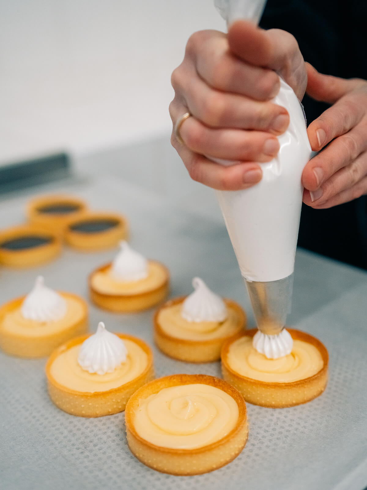 Aus einem Spritzbeutel mit feiner Sterntülle wird ein weißer Baisertupfen mittig auf ein mit heller Zitronencreme gefülltes Tartelette gesetzt