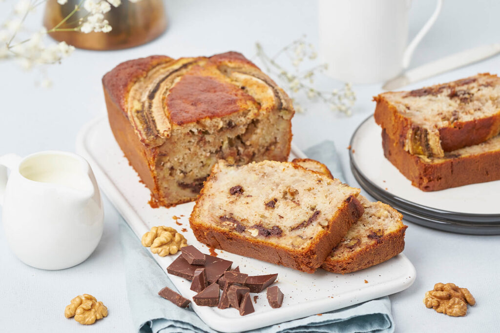 Angeschnittenes Bananenbrot mit Walnüssen und Schokolade auf einer weißen Platte neben einem Milchkännchen