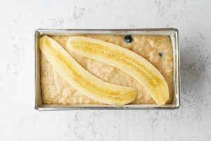 Bananenteig mit Heidelbeeren in der gefetteten Kastenform, belegt mit zwei längs halbierten Bananen