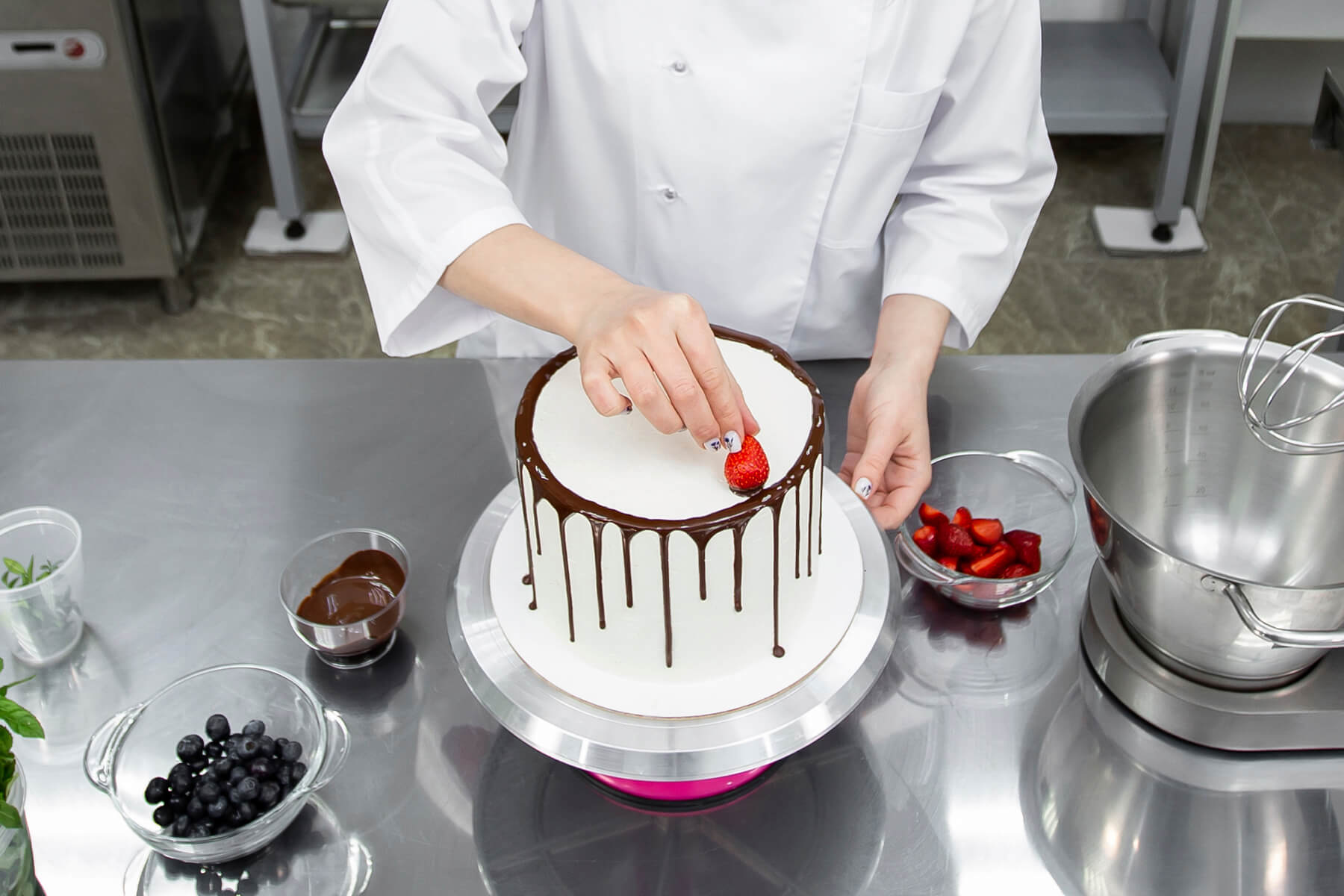 Blick von oben auf eine Torte mit Schokoladenrand, daneben Schalen mit geschnittenen Erdbeeren, Heidelbeeren und geschmolzener Schokolade