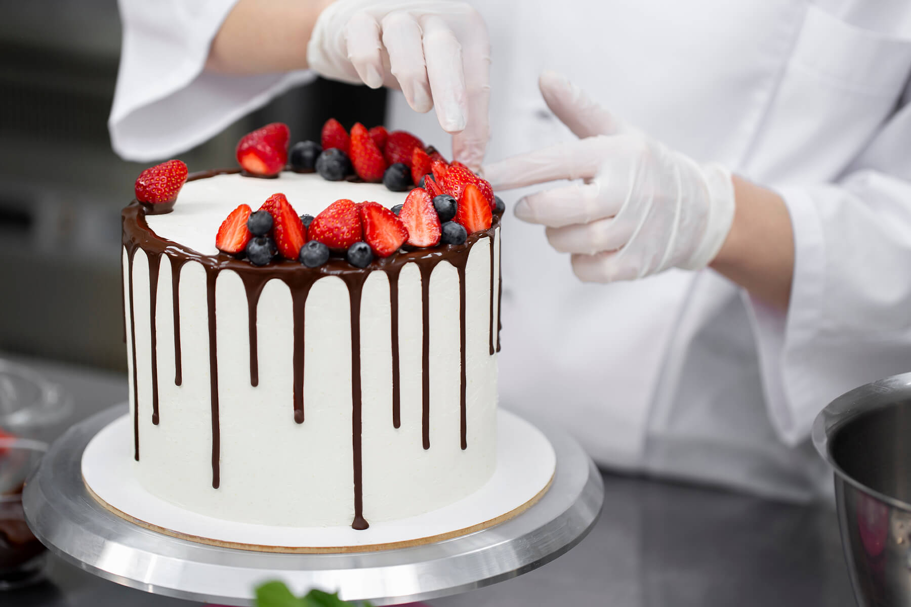 Hände in Einweghandschuhen ordnen Erdbeeren und Heidelbeeren auf einer weißen Torte mit Schokoladenrand