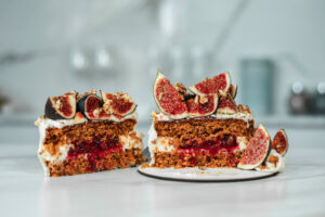 Zwei Hälften der angeschnittenen kleinen Torte nebeneinander, im Schnitt sind zwei Möhrenkuchenböden, weiße Creme und eine rote Beerenfüllung zu sehen, obenauf Feigenviertel und gehackte Walnüsse