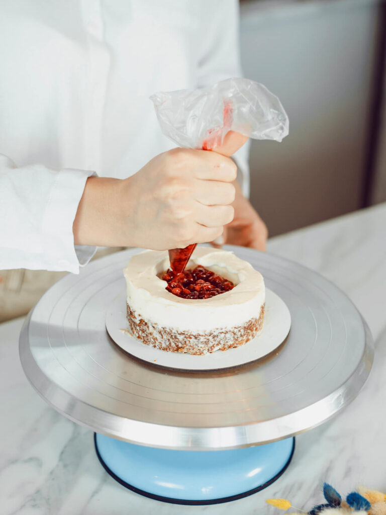 Rote Beerenfüllung wird aus einem Spritzbeutel in die Mitte des weißen Cremekranzes auf der kleinen Torte gefüllt