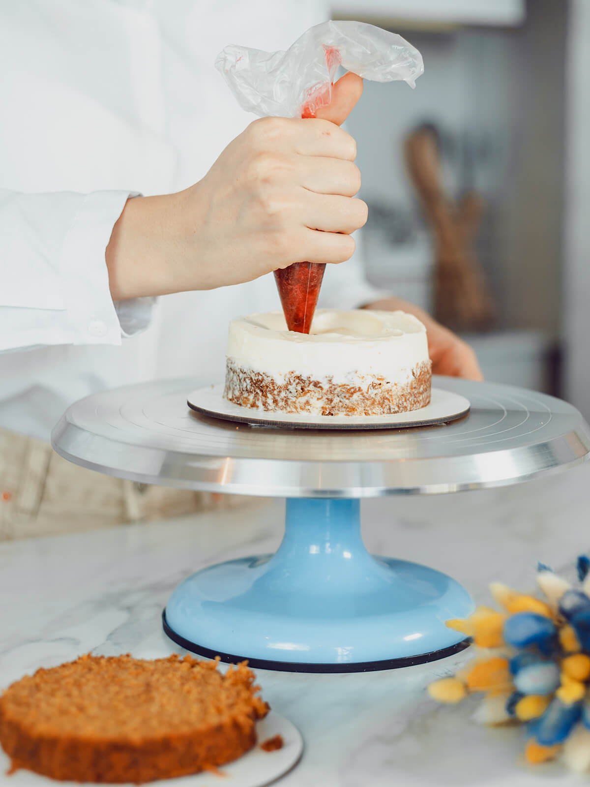Roter Beerenfüllung wird aus einem Spritzbeutel in die Vertiefung des weißen Cremewalls auf der kleinen Torte gefüllt, der zweite Boden liegt im Vordergrund