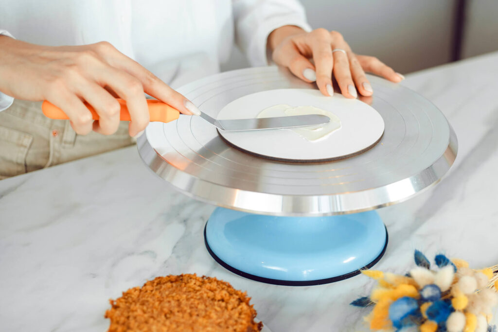 Hand mit oranger Winkelpalette verstreicht einen Klecks weiße Creme auf einer runden weißen Cakeboard-Scheibe auf einem Drehteller