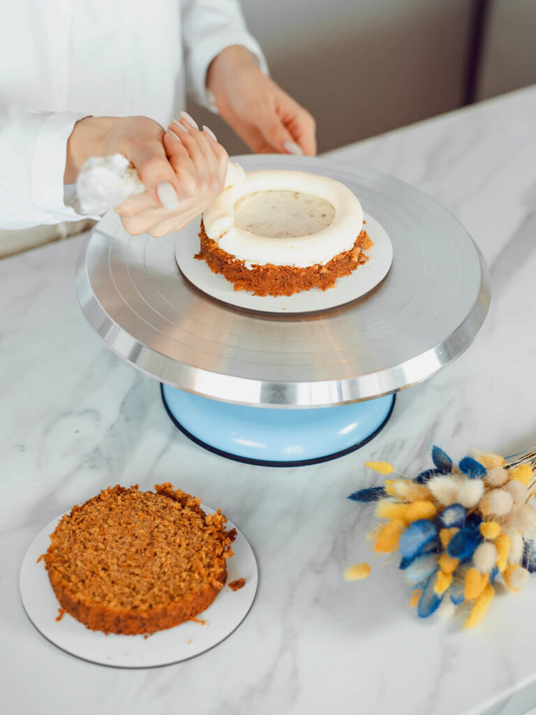 Hand spritzt mit einem Spritzbeutel einen dicken weißen Cremekranz auf den Rand eines Möhrenkuchenbodens, die Mitte ist dünn mit Creme bestrichen, der zweite Boden liegt daneben