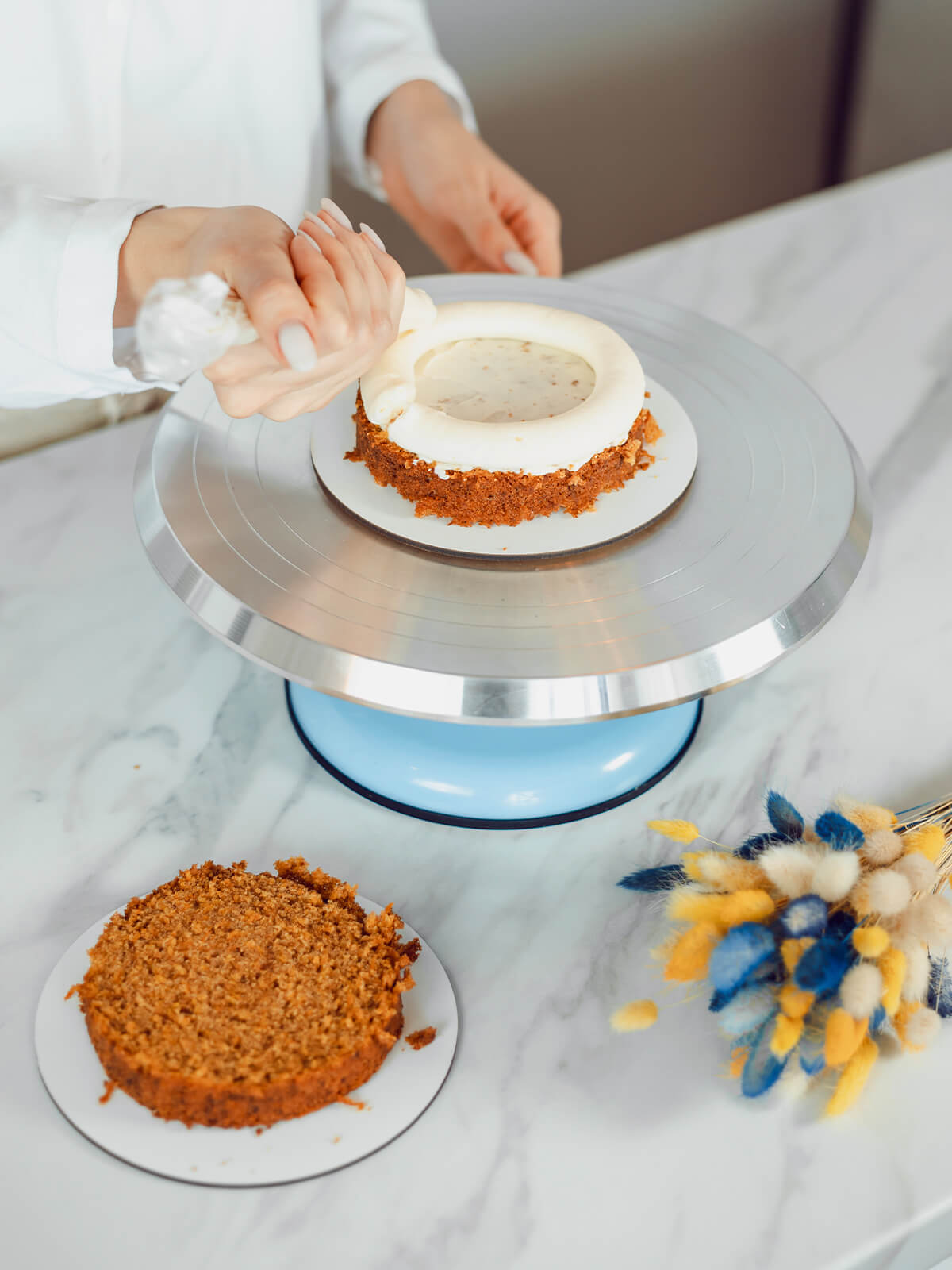 Hand spritzt mit einem Spritzbeutel einen dicken weißen Cremekranz auf den Rand eines Möhrenkuchenbodens, die Mitte ist dünn mit Creme bestrichen, der zweite Boden liegt daneben