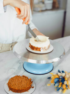 Hand formt mit einer Winkelpalette den hohen weißen Cremewall auf dem Möhrenkuchenboden, in der Mitte ist eine deutliche Vertiefung entstanden