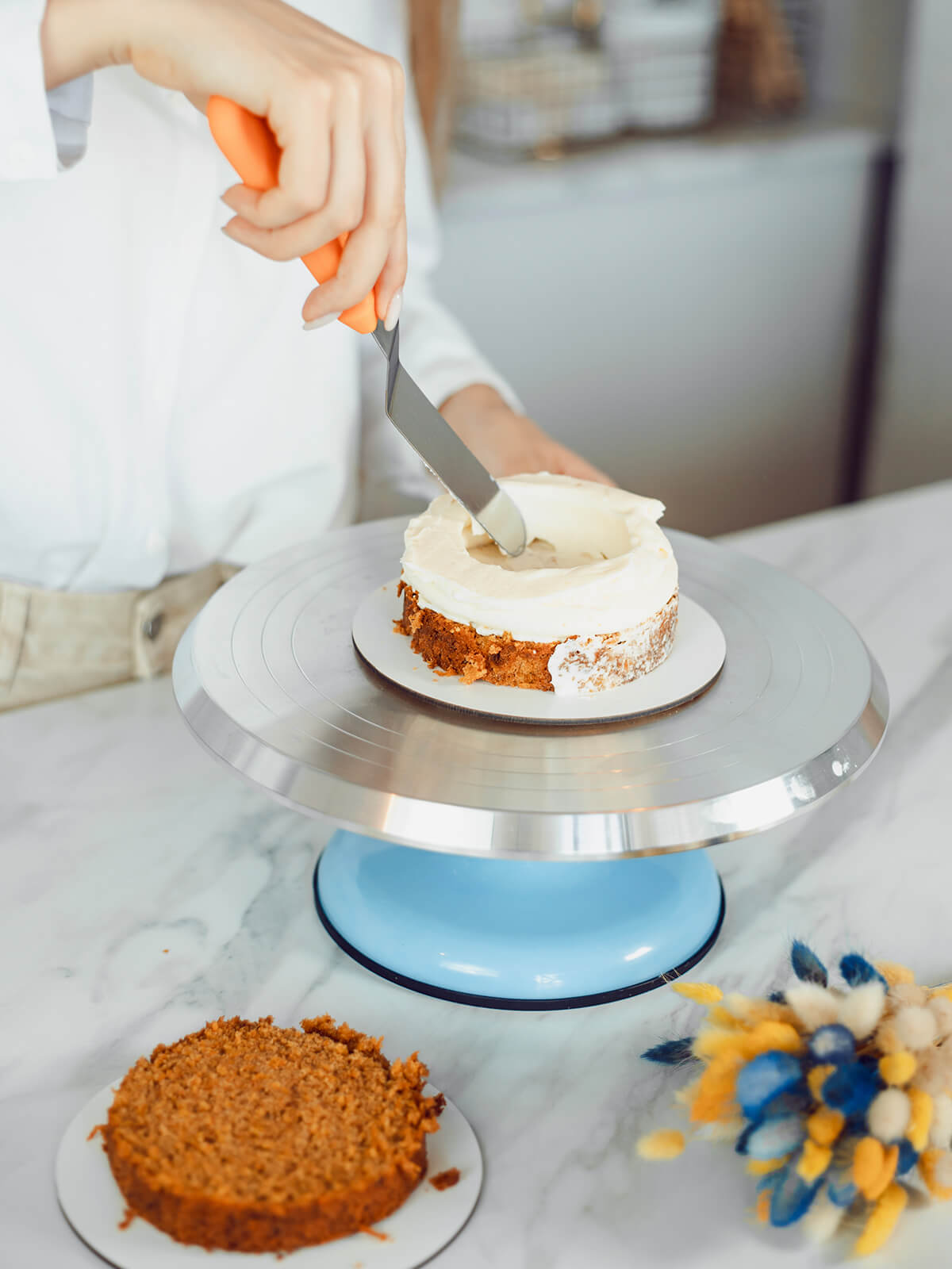 Hand formt mit einer Winkelpalette den hohen weißen Cremewall auf dem Möhrenkuchenboden, in der Mitte ist eine deutliche Vertiefung entstanden