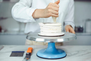 Weiße Creme wird in dicken, durchgehenden Ringen von unten nach oben um eine zweistöckige Torte gespritzt, eine Teigkarte liegt bereit
