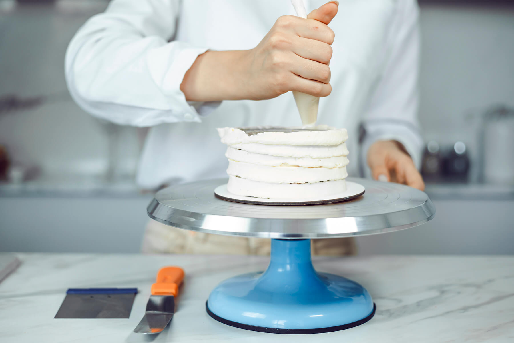 Weiße Creme wird in dicken, durchgehenden Ringen von unten nach oben um eine zweistöckige Torte gespritzt, eine Teigkarte liegt bereit