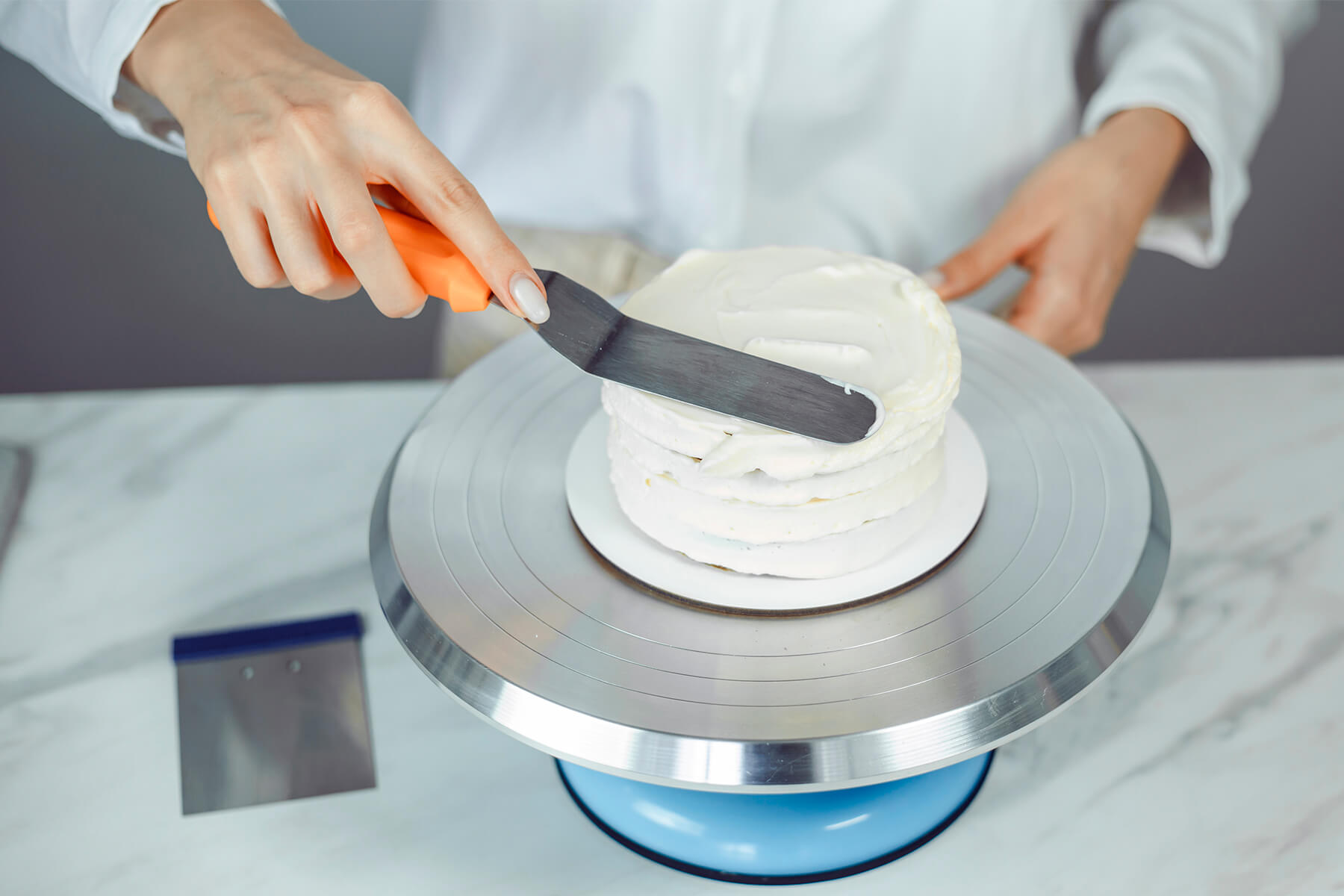 Winkelpalette zieht die weiße Creme auf der Oberseite einer kleinen Torte glatt, die Seiten sind noch in Ringen aufgespritzt, eine Teigkarte liegt bereit