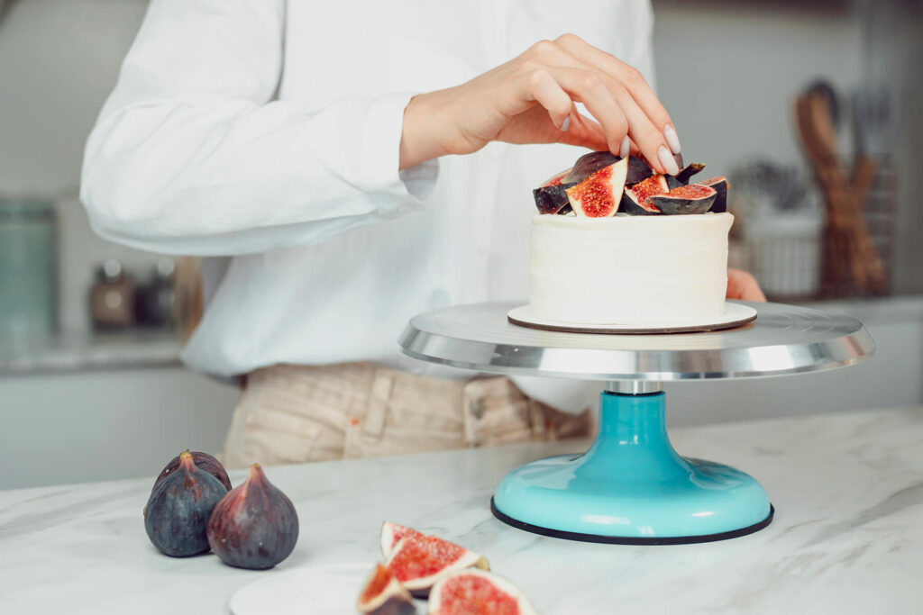 Hand setzt das letzte Feigenviertel auf den fertigen Feigenkranz der Bento-Torte, die auf einem türkisfarbenen Tortenständer steht