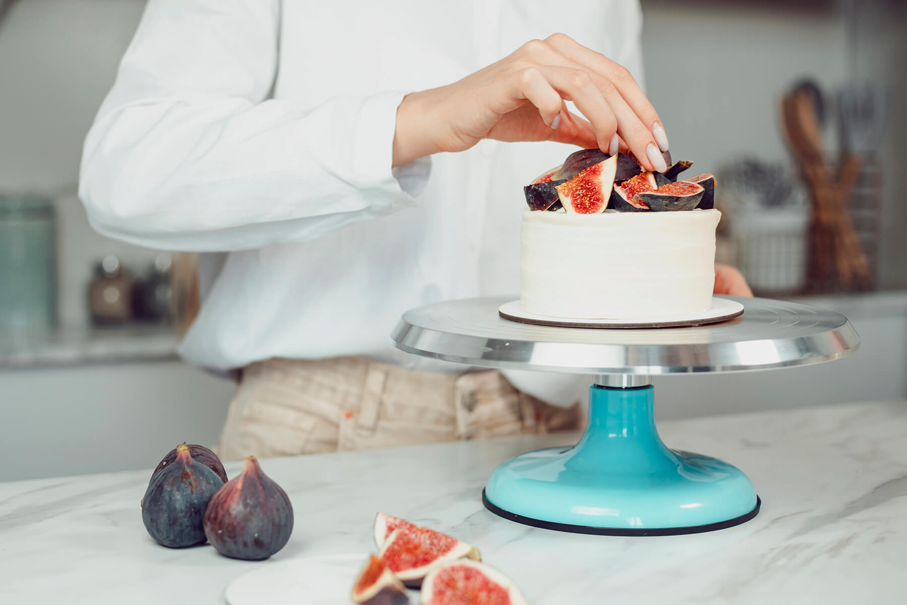 Hand setzt das letzte Feigenviertel auf den fertigen Feigenkranz der Bento-Torte, die auf einem türkisfarbenen Tortenständer steht