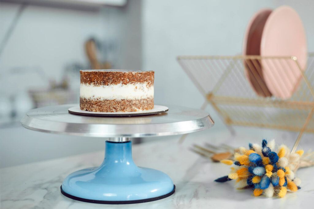 Kleine Torte mit dünner, gestreifter Krümelschicht auf einem Drehteller, die Möhrenböden sind durch die Creme hindurch sichtbar