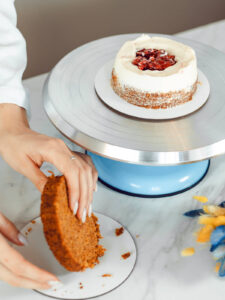 Hände heben einen Möhrenkuchenboden an, im Hintergrund steht die kleine Torte mit rotem Beerenkern im weißen Cremekranz auf dem Drehteller