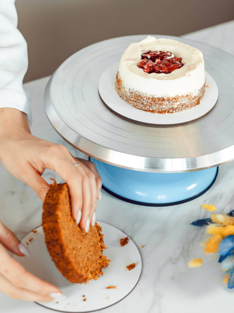 Hände heben einen Möhrenkuchenboden an, im Hintergrund steht die kleine Torte mit rotem Beerenkern im weißen Cremekranz auf dem Drehteller
