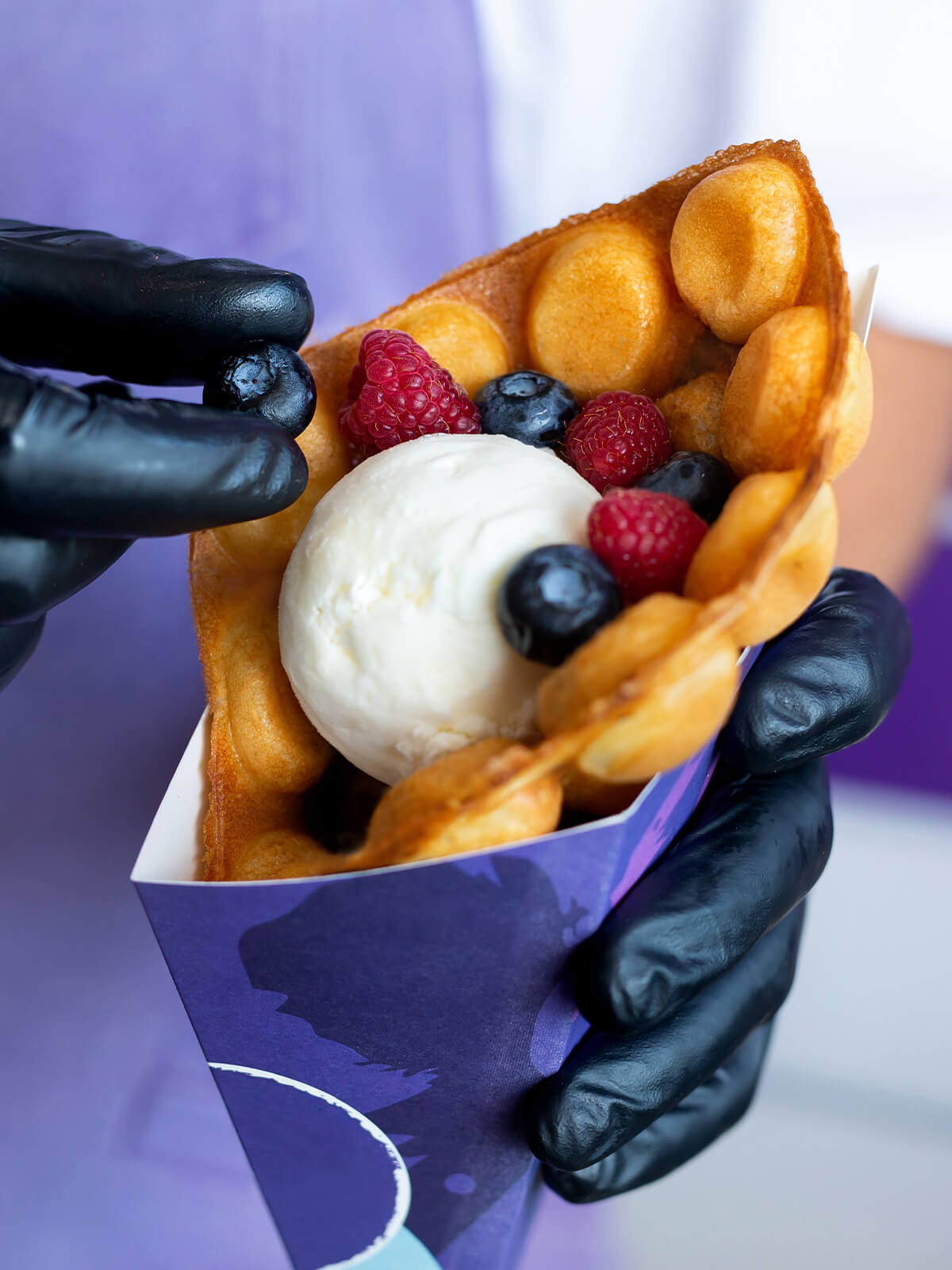 Zur Tüte gerollte Bubble Waffle im Papierhalter, gefüllt mit einer Kugel Eis, Himbeeren und Heidelbeeren
