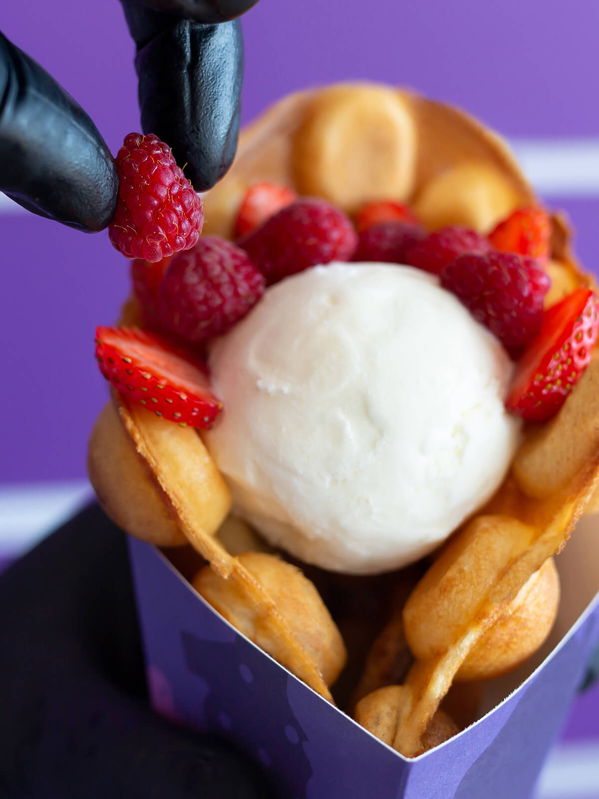 Eine behandschuhte Hand legt eine Himbeere auf die Eiskugel in der gefalteten Bubble Waffle