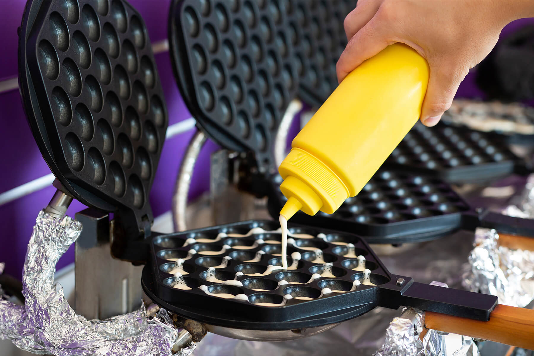 Aus einer gelben Dosierflasche läuft dünner Waffelteig in die halbrunden Mulden eines heißen Bubble-Waffle-Eisens
